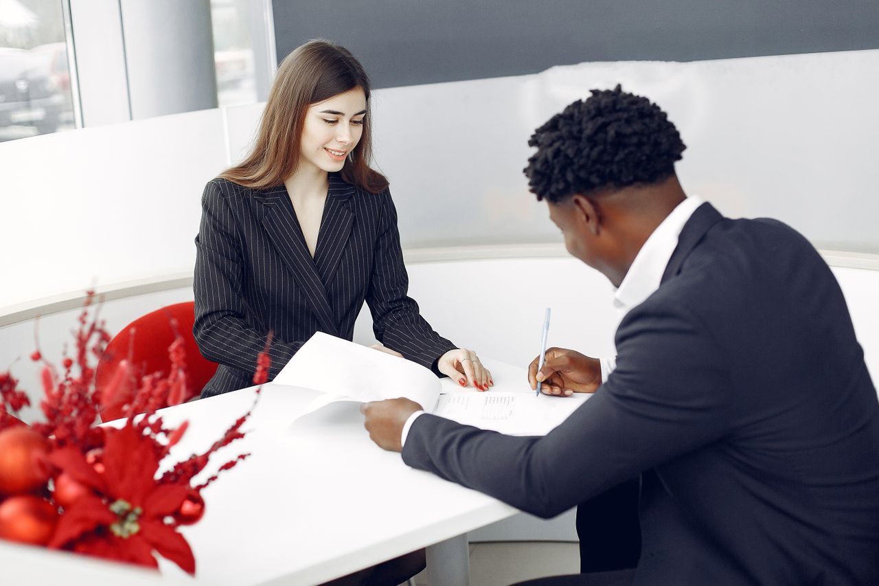 our-story Positive stylish multiethnic buyer in formal classy suit and agent signing papers in modern light creative office in daylight and smiling happily